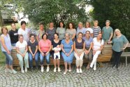 19 Studierende mit Schulleiterin sitzen auf einer Holzbank vor dem Schulgebäude.