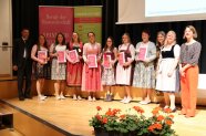 11 Personen stehen auf einer Bühne, davon die 7 Absolventinnen mit Zeugnissen in den Händen